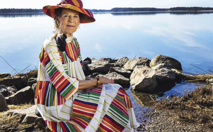 Värikkääseen hameeseen pukeutunut nainen istuu kalliolla järven rannalla. Taustalla näkyy tyyni järvi ja sininen taivas. Lehtikuvan otsikko on 'Järvenpääläinen KESÄ 1 2026' ja siinä mainitaan 'Järvenpää 75 vuotta'.