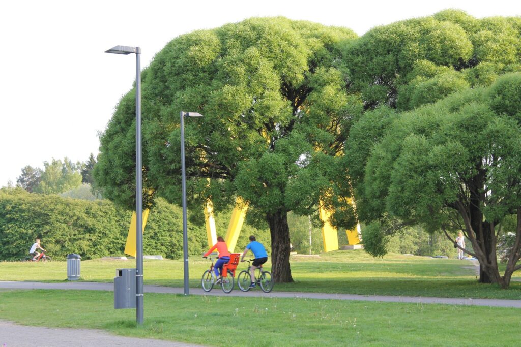 Kaksi pyöräilijää ajaa puistossa asfaltilla. Heidän taustallaan on suuria, vehreitä puita, joiden oksista roikkuu keltaisia lippuja.