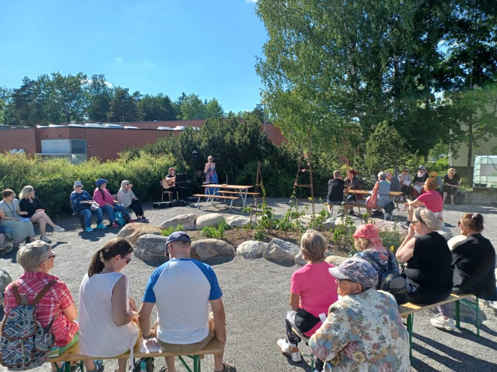 Ihmisiä istumassa puolikaaressa Kesälaitumen kentällä, kuuntelemassa musiikkiesitystä.