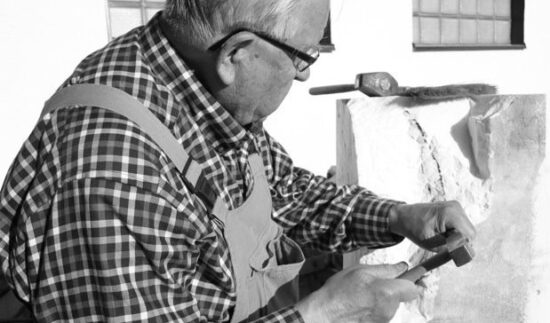 Iäkäs mies työstää vaaleaa marmoriveistosta työpajassaan. Hän nojaa keskittyneenä orgaanisen muotoiseen Carraran marmorista veistettyyn teokseen, ja etualalla näkyy kiviveistotyökaluja.