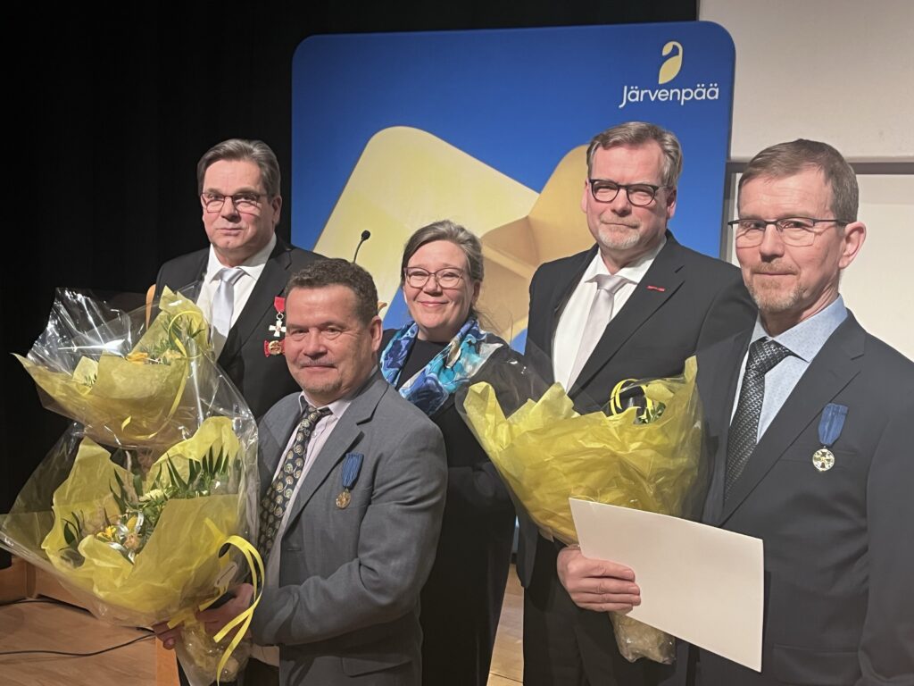 Viisi henkilöä seisoo kuvassa ja kolmella henkilöllä on keltasävytteiset kukkakimput kädessä.