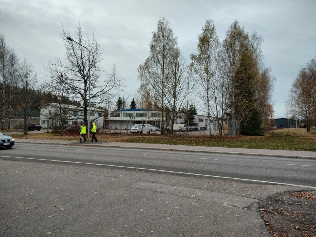 Asfalttitietä, puita ja yritysrakennus aitojen takana. Ihmisiä kävelee kadulla.