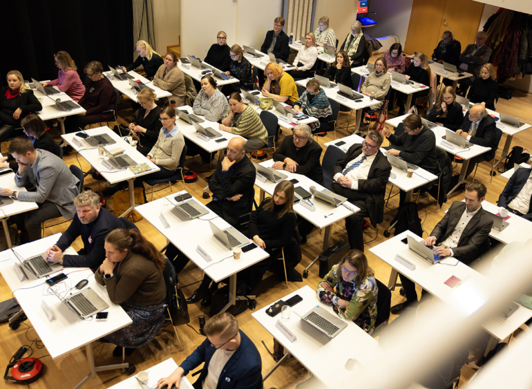 Ihmiset istuvat kokoustilassa, jossa on riveissä valkoisia pöytiä ja kannettavia tietokoneita. He näyttävät keskittyvän edessä tapahtuvaan esitykseen.