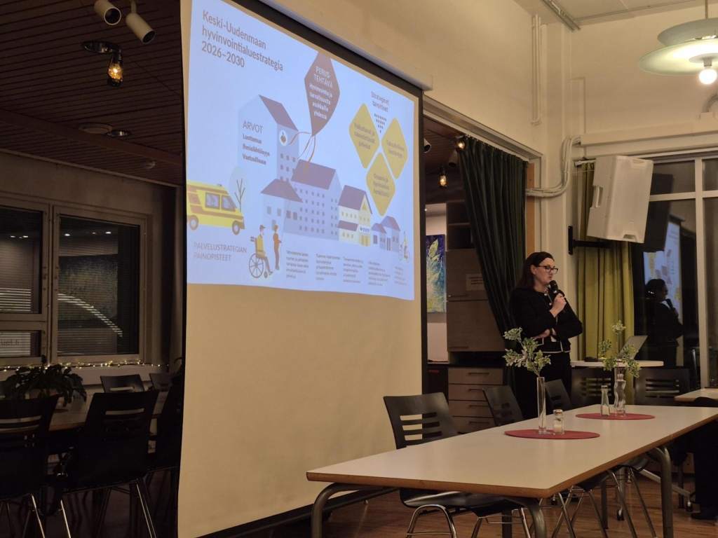 Henkilö esittelee strategiaa projektorilla valaistulla valkokankaalla sisätilassa. Valkokankaalla lukee 'Keski-Uudenmaan hyvinvointialuestrategia 2026–2030' ja siinä on kaavioita ja tekstiä.