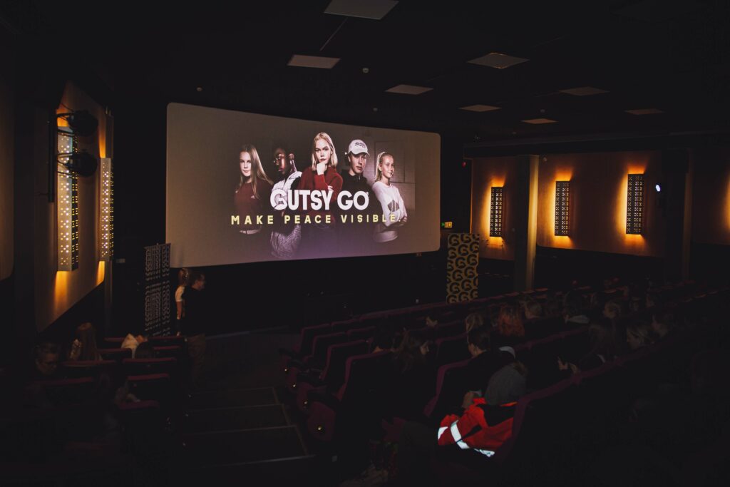 Elokuvateatterin valkokankaalle heijastettu näkymä, jossa nuoria henkilöitä ja keskellä teksti "Gutsy Go: Make Peace Visible".