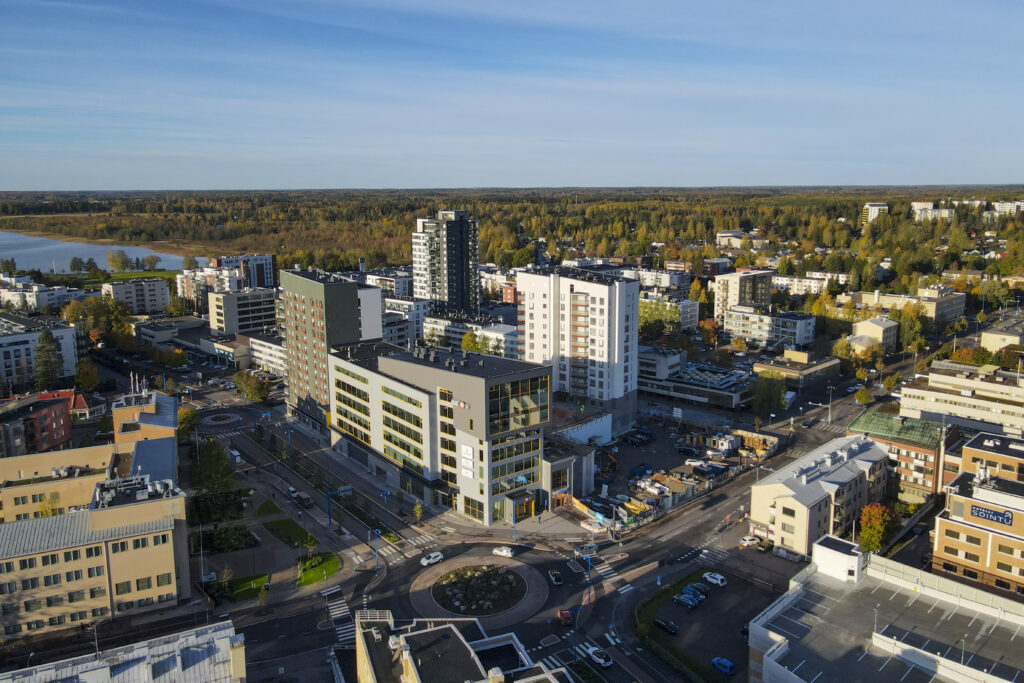 Ilmakuva Järvenpään kaupungista, jossa näkyy moderneja kerrostaloja, liikekeskuksia ja liikenneympyrä etualalla. Taustalla avautuu metsämaisema ja järvi, joka korostaa alueen vehreyttä ja luonnonläheisyyttä.