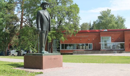 Jean Sibeliuksen pronssinen patsas seisoo nurmialueella Järvenpään taidemuseon edustalla. Taustalla näkyvät museorakennus, lehtipuita ja kesäinen sinitaivas.