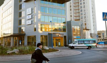Järvenpään kaupungin toimitalo Bulevardikorttelissa syysauringossa. Etualalla kävelijä potkulaudan kanssa ja kaupungin minibussi Järvenpää-kuosilla kääntymässä liikenneympyrässä. Rakennuksen julkisivussa näkyvät Järvenpään kaupungin ja muiden toimijoiden kyltit.