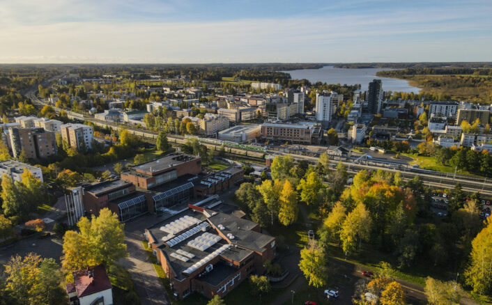 Ilmakuva Järvenpään keskustasta syksyisessä maisemassa. Kuvassa näkyy rautatieasema, asuinkerrostaloja, kaupungin rakennuksia sekä Tuusulanjärvi taustalla. Etualalla on punatiilisiä rakennuksia ja värikkäitä syyspuita.