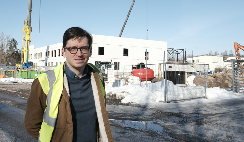 Eero Renell seisoo kuvan etuosassa huomioliivit päällään, taustalla vaalea rakenteilla oleva koulurakennus.