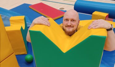 Mies istumassa sisätemppuradan välineiden seassa. Taustalla näkyy sählyä pelaavia poikia.