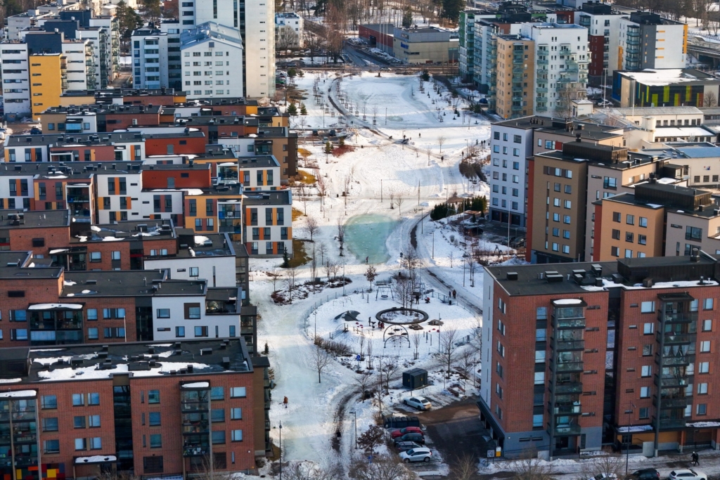 Ilmakuva talvisesta asuinalueesta, jossa kerrostalot ympäröivät lumista puistoaluetta. Puiston halki kulkee kävelyreittejä, keskellä näkyy pieni osittain jäätynyt lampi ja leikkipaikka.