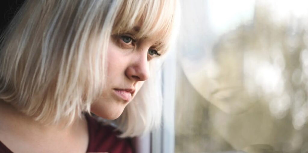 Nainen katsoo mietteliäänä ulos ikkunasta, hänen heijastuksensa näkyy lasissa.