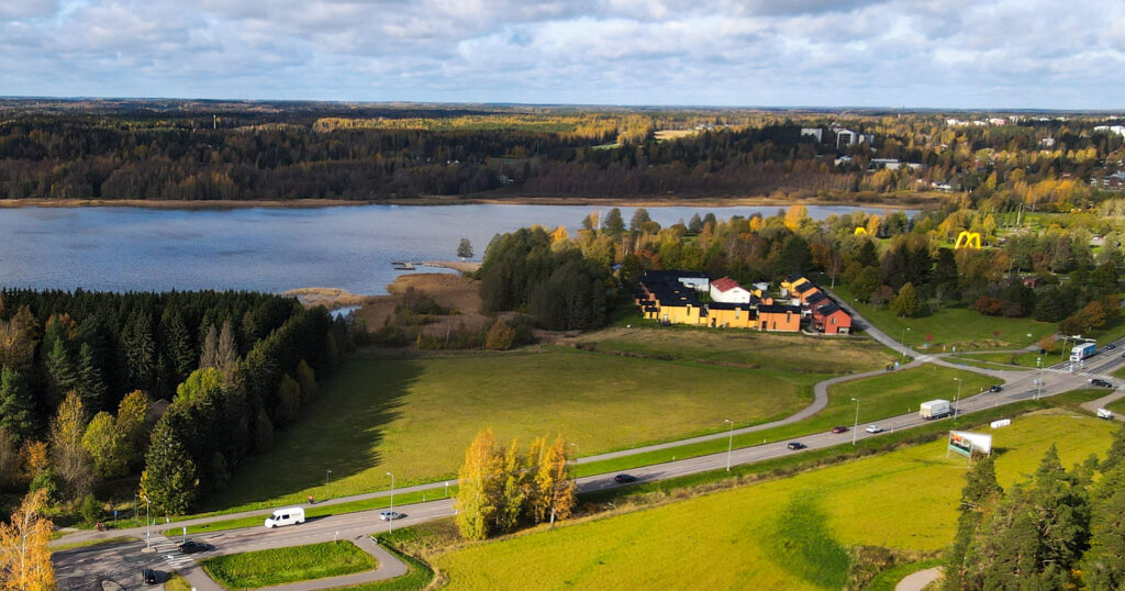 Ilmakuva Järvenpäästä, etualalla vihreitä peltoja ja liikenneväyliä, taustalla järvi ja metsää. Kuvassa näkyy myös rakennuksia ja syksyisiä puita keltaisine ja ruskeine lehtineen.