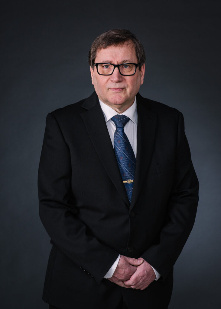 Mies mustassa puvussa ja sinisessä solmiossa ja hänellä on silmälasit.