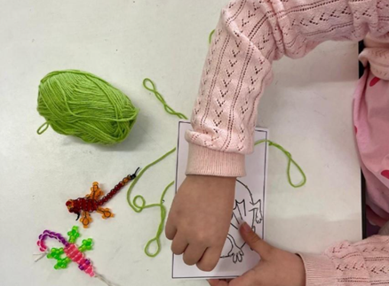 Ylhäältä päin otettu kuva, jossa näkyy lapsen kädet. Lapsi tekee ompelutyötä paperiin. Vieressä on vihreä lankakerä.