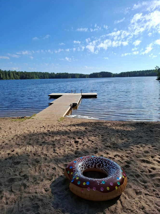 Hiekkaranta, jossa laituri ja hiekalla donitsin muotoinen uimarengas. Taustalla järvimaisema.