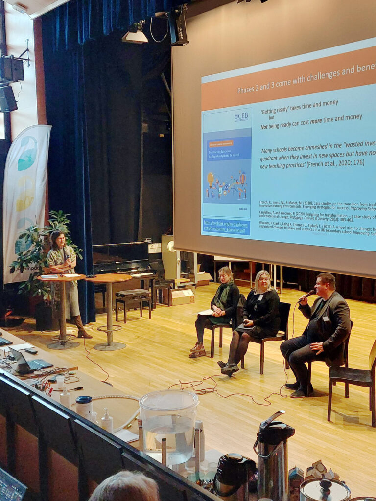 Järvenpään lukion auditorion lavala kolme neljä henkilöä keskustelemassa. Taustalla suuri näyttökangas.