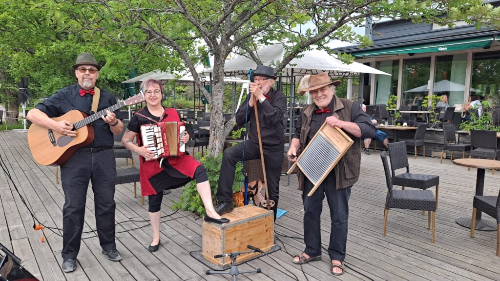 Tuusulanjärven Skiffle-orkesteri paikallisen ravintolan terassilla. Orkesterin neljä jäsentä ja soittiminaan kitara, haitari, laulu ja pyykkiteline.