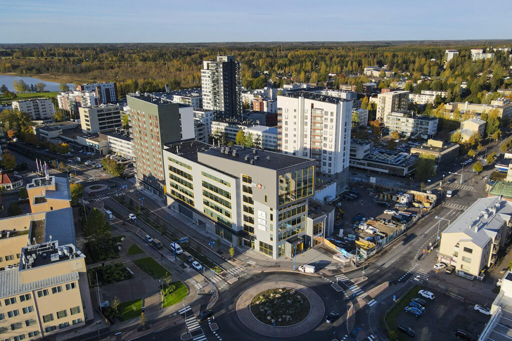 Ilmakuva kaupunkimaisemasta, jossa on useita kerrostaloja, pyöreä liikenneympyrä ja taustalla näkyy metsää ja järvi.