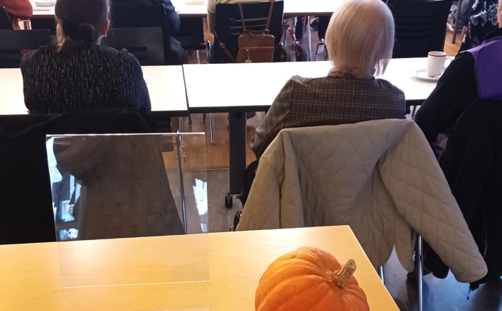 Ihmiset istuvat luokkahuoneessa tai kokoushuoneessa kuuntelemassa ja keskustelemassa. Etualalla pöydällä on kolme pientä kurpitsaa.