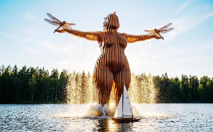 Puinen, suuri naisfiguuria esittävä veistos seisoo järvessä kädet levitettyinä; käsistä roikkuu vettä ja niiden päissä on sudenkorennon siivet. Taustalla metsäinen rantaviiva ja veistoksen edessä pieni purjevene.