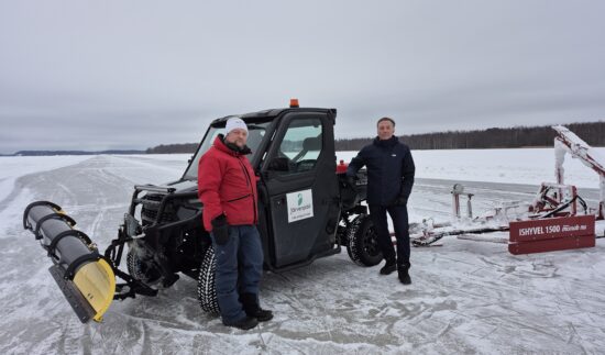 Kaksi miestä seisoo järven jäällä. Jäällä on myös koppimönkijä, jonka perässä jäähöylä. Taustalla näkyy järvellä olevaa jäärataa.