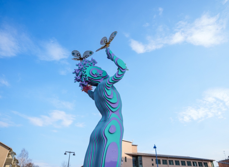 Värikäs Järvenhaltija-veistos Järvenpäässä pitelee käsissään sudenkorentoja sinistä taivasta vasten.