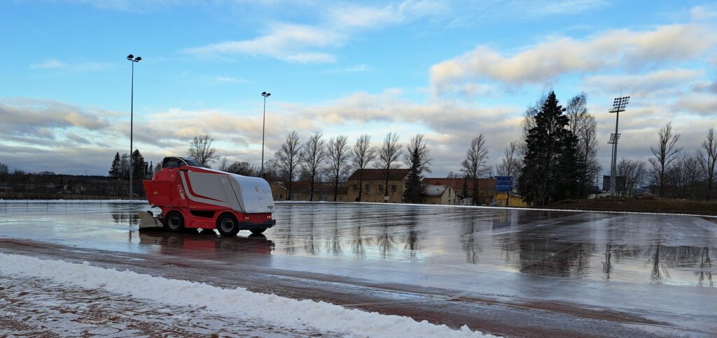 Luistelukenttä, jolla on jääkone huoltamassa jääpintaa kirkkaana talvipäivänä.