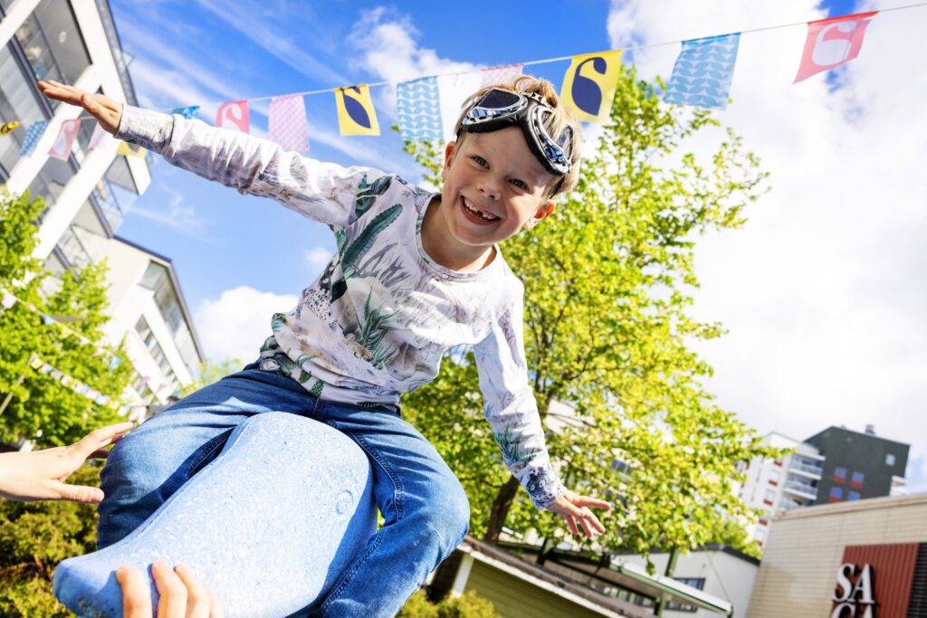 Kuvassa iloinen lapsi on patsaan päällä lentäjän asennossa otsallaan lentäjän lasit.