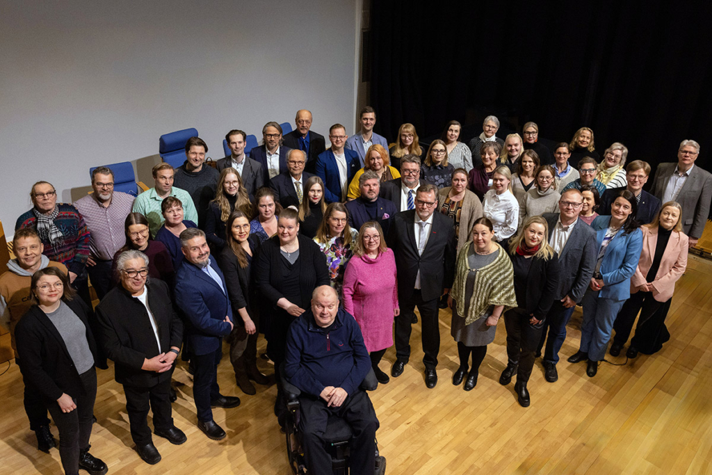 Ryhmä ihmisiä seisoo sisätilassa, useat hymyilevät kameralle. Eturivissä on henkilö pyörätuolissa.