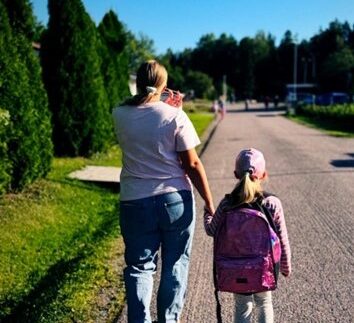 Vanhempi ja lapsi kävelevät käsi kädessä tiellä. Lapsella on reppu selässä.