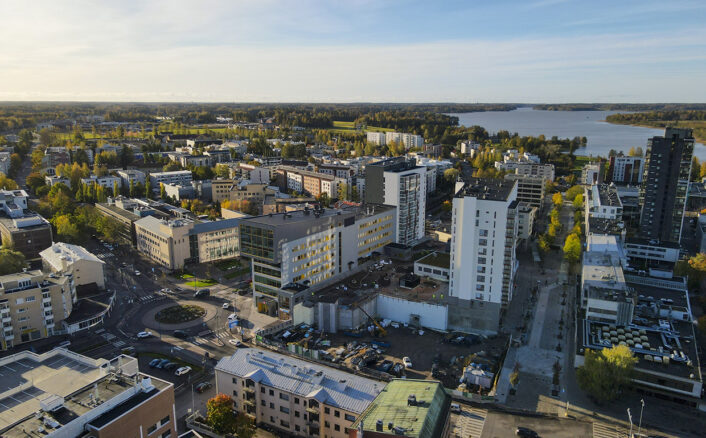 Ilmakuva kaupungista, jossa on useita korkeita rakennuksia ja vehreitä alueita. Taustalla näkyy järvi ja metsä.
