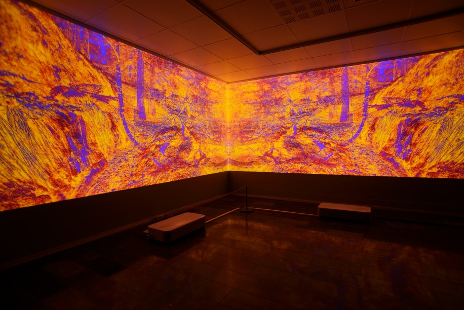 Huoneen seinille projisoitu värikäs taideteos, joka näyttää abstrakteja oranssin ja sinisen sävyjä. Teos luo voimakkaan visuaalisen vaikutelman pimeässä tilassa.