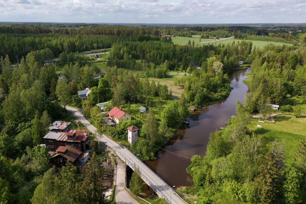 Ilmakuva vehreästä jokimaisemasta maaseudulla. Kuvassa näkyy mutkitteleva joki, sitä ylittävä pieni silta sekä vanhoja rakennuksia ja muutamia asuintaloja. Ympärillä on runsaasti puita ja peltoja, ja taustalla avautuu metsäinen maisema.