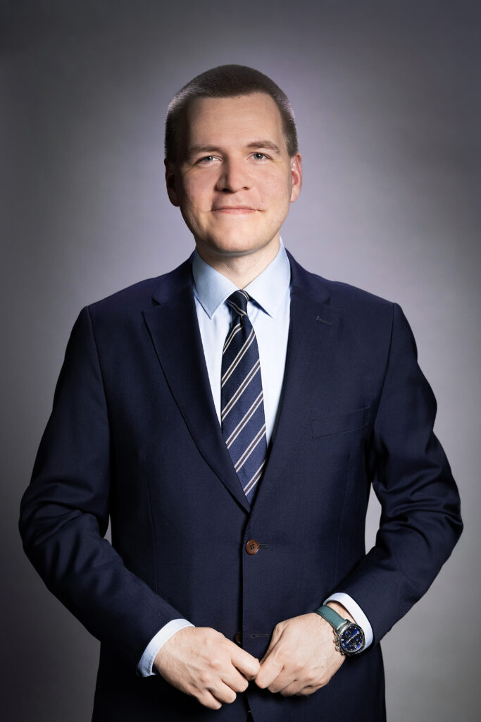 Henkilö pukeutunut tummansiniseen pukuun ja raidalliseen solmioon, katsoo kameraan ja hymyilee.