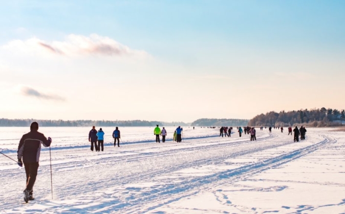 Ihmiset ulkoilevat talvisella, lumen peittämällä jäällä; etualalla henkilö liikkuu sauvojen kanssa, taustalla useita kävelijöitä ja luistelijoita avaralla järvimaisemalla kirkkaassa talvisäässä.