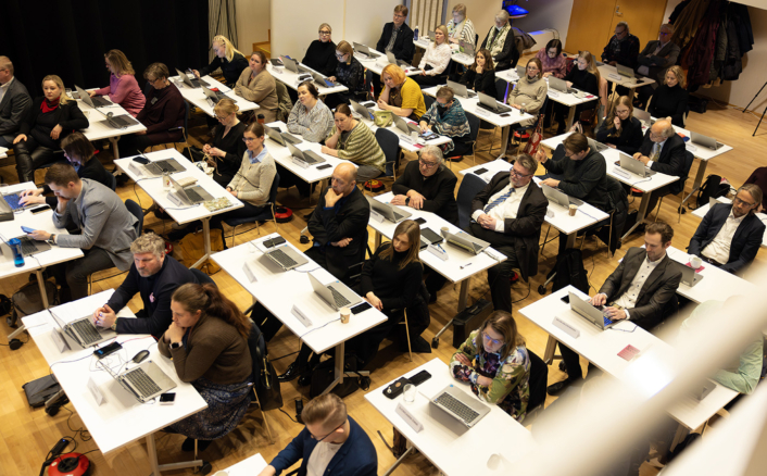 Ihmiset istuvat kokoustilassa, jossa on riveissä valkoisia pöytiä ja kannettavia tietokoneita. He näyttävät keskittyvän edessä tapahtuvaan esitykseen.