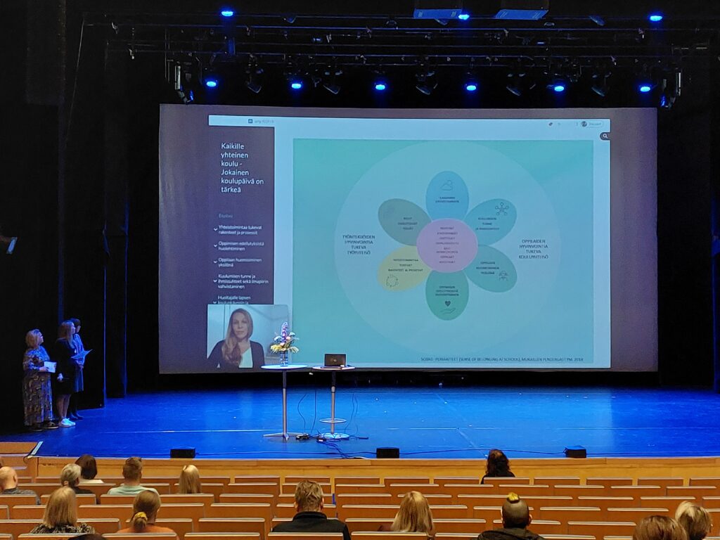 Juhlatilan lavalla esitys, jossa värikkäässä kuviossa tekstiä. Lavalla reunalla ihmiset katsovat esityksen suuntaan.