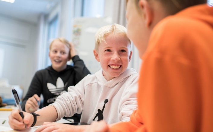 Kolme lasta istuu pöydän ääressä luokkahuoneessa. Keskellä oleva lapsi hymyilee ja pitää kynää kädessä. Lapsilla on päällään hupparit, ja he näyttävät olevan hyvällä tuulella.