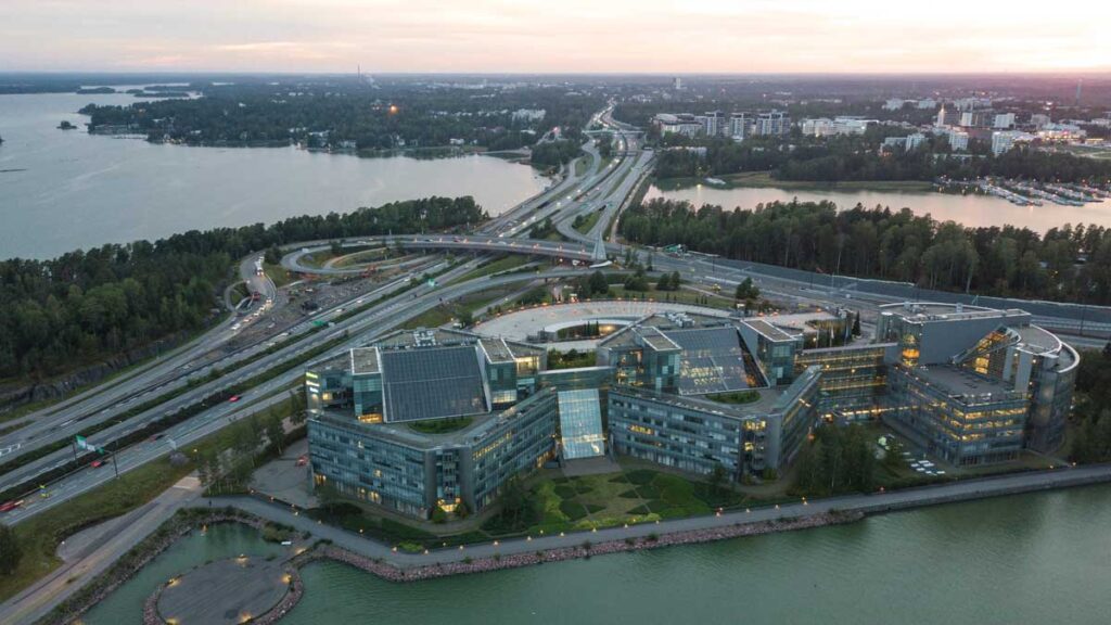 Tämä on ilmakuva Espoosta, jossa näkyy Keilaniemen toimistoalue ja sen rannassa sijaitseva näyttävä toimistokompleksi meren äärellä. Kuvan etualalla näkyvä rakennus on valmistunut modernilla arkkitehtuurilla ja sijaitsee aivan veden rajassa, taustalla kulkevat Länsiväylä ja Kehä I, jotka johtavat kohti Helsingin keskustaa. Kuvassa näkyy myös Espoon rantaraittia, venevalkamia ja metsien reunustamia asuinalueita ilta-auringon valossa.