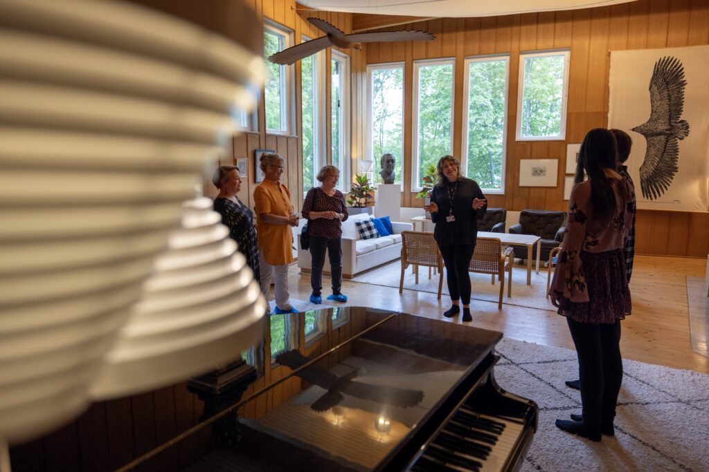 Ryhmä ihmisiä seisoo puupaneelisessa olohuoneessa, jossa on suuret ikkunat ja sisustuselementtejä, kuten flyygeli, sohva ja taideteos seinällä.