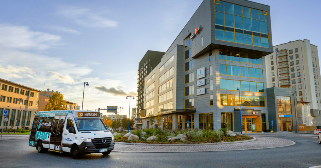 Bussi pysähtyy nykyaikaisen liikerakennuksen edessä, ilta-auringon valaistessa ympäristöä.