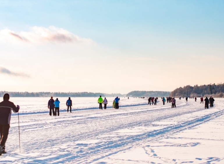Ihmiset ulkoilevat talvisella, lumen peittämällä jäällä; etualalla henkilö liikkuu sauvojen kanssa, taustalla useita kävelijöitä ja luistelijoita avaralla järvimaisemalla kirkkaassa talvisäässä.