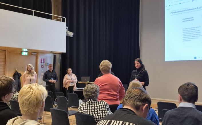 Ihmisiä istumassa ja seisomassa auditoriossa, jossa pidetään esitystä. Seinällä on suuri valkokangas, johon on projisoitu tekstiä. Nainen seisoo edessä puhumassa.