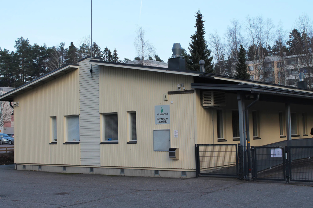 Järvenpään Perhetalo Joutsikin vaalea yksikerroksinen rakennus Naavatien varrella. Rakennuksen sisäänkäynnin vieressä on Perhetalo Joutsikin kyltti, ja piha-alue on aidattu. Taustalla näkyy puita ja kerrostaloja.
