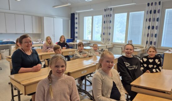 Oppilaat ja yksi aikuinen henkilö istuvat pulpeteissa luokkahuoneessa. He näyttävät iloisilta.