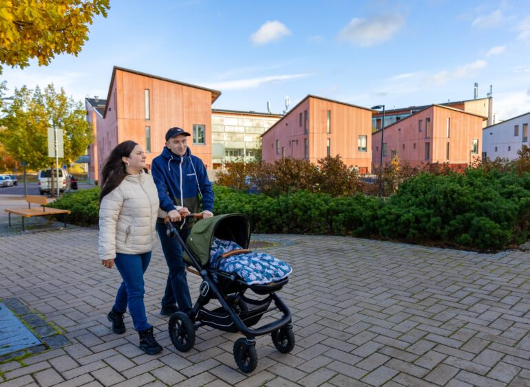 Pariskunta kävelee ulkona rattaiden kanssa modernien punaisten rakennusten edessä.