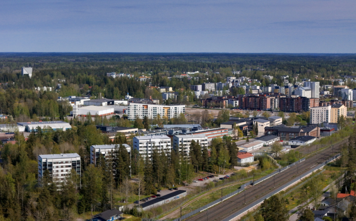 Ilmakuva Järvenpään kaupunkimaisemasta: etualalla kulkee rautatie rata-alueineen, sen ympärillä pientaloja ja kerrostaloja. Taustalla näkyy laaja asuinalue, matalia ja keskikorkeita rakennuksia sekä runsaasti vihreää metsää ja puustoa, joka ympäröi kaupunkia. Horisontissa avautuu tasainen metsämaisema kirkkaan taivaan alla.