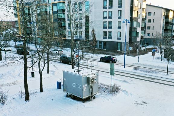 Kaupungin katunäkymää, jossa ajaa autoja. Taustalla kerrostaloja. Edustalla on ilmanlaadun mittausasema.